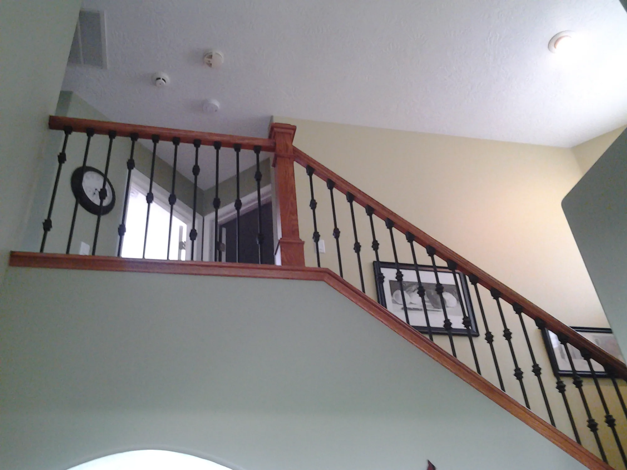Stained wood handrail running up the rake of a stair and returning level along an upper landing, with black iron balusters between a top rail and a shoe rail and a square newel post at the turn
